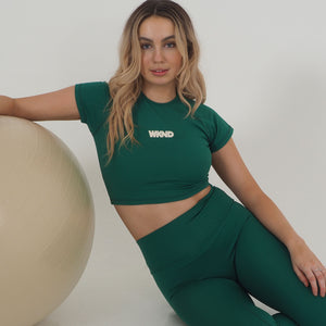 Model with blonde hair wearing forest green leggings and matching crop top. Model is sitting on the floor and leaning on a yoga ball.