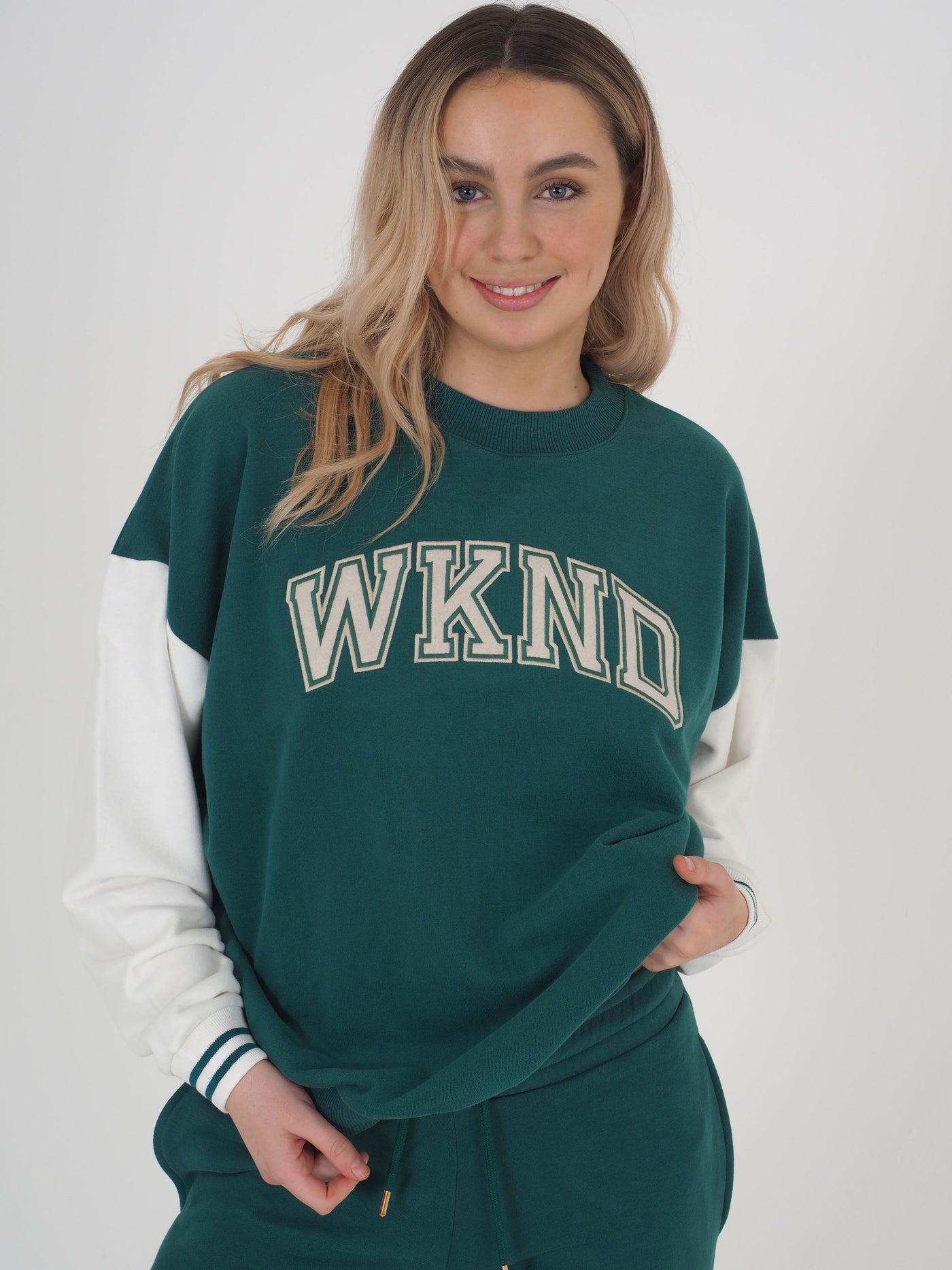 Model with blonde hair wearing a green college varsity style sweatshirt with matching joggers..  Sweatshirt has contrast white sleeves and the text reads WKND.