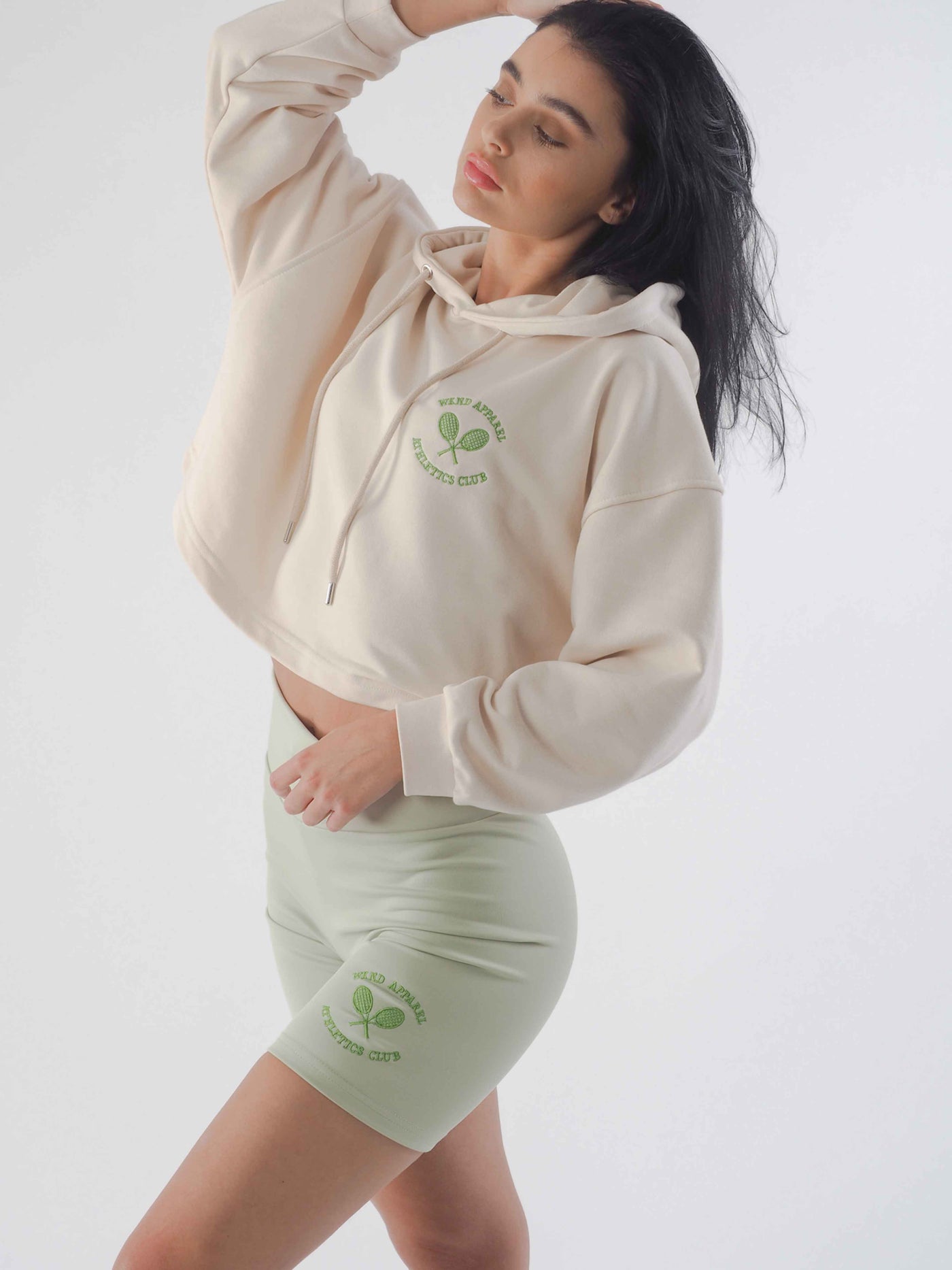 Model is posing in a cropped hoodie and cycling shorts in jade green. Both products have matching WKND Apparel embroidery.