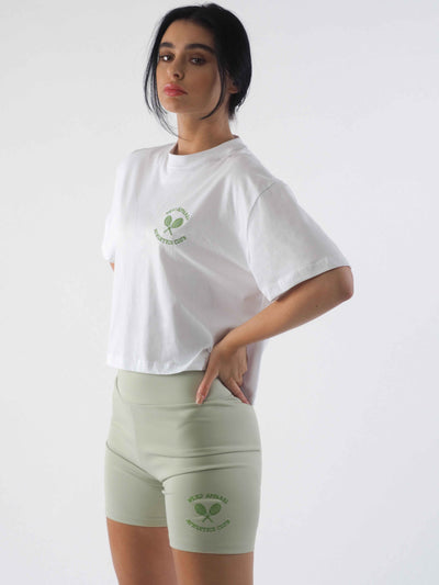 Model with dark hair tied up, wearing an oversized cropped white t-shirt and jade green cycling shorts. The WKND Apparel tennis logo is embroidered to the chest and the leg.