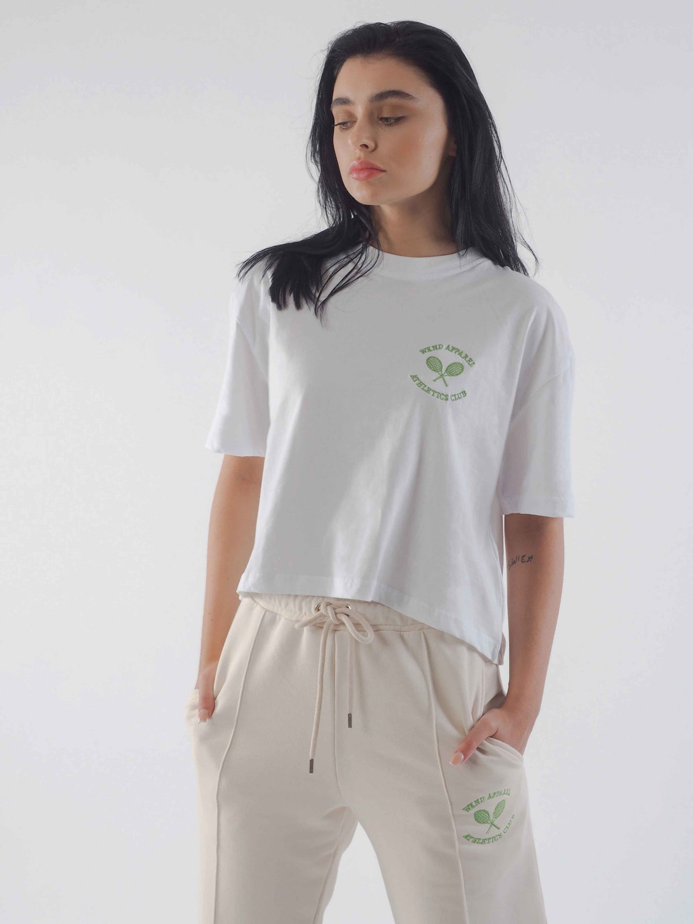 Model with dark hair wearing an oversized cropped white t-shirt. The WKND Apparel tennis logo is embroidered to the chest.  Model is also wearing eggnog joggers.