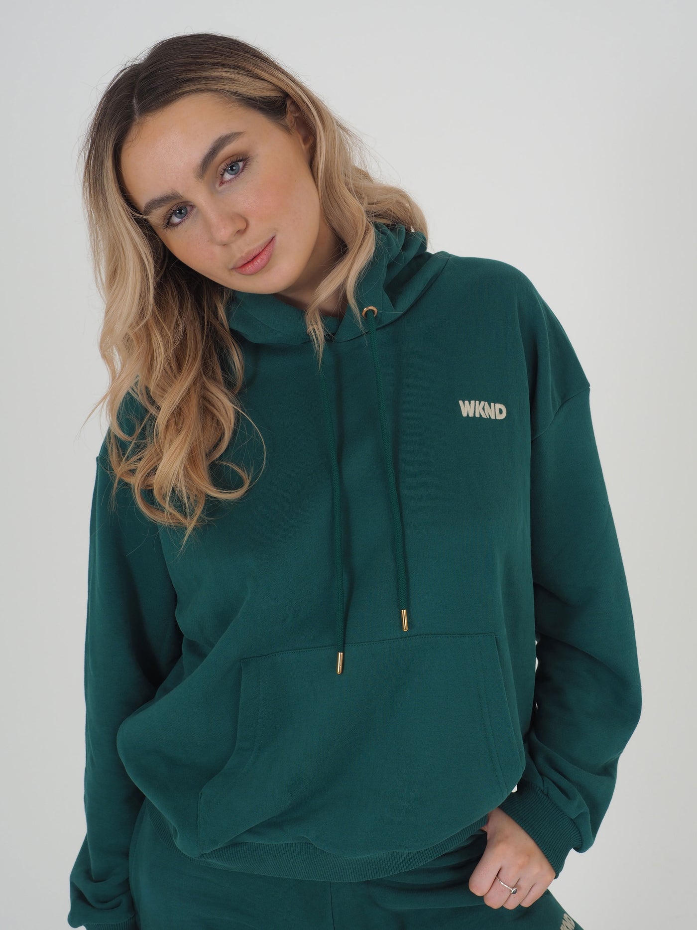 Model with blonde hair is wearing a green hoodie. WKND Embroidery to the chest.