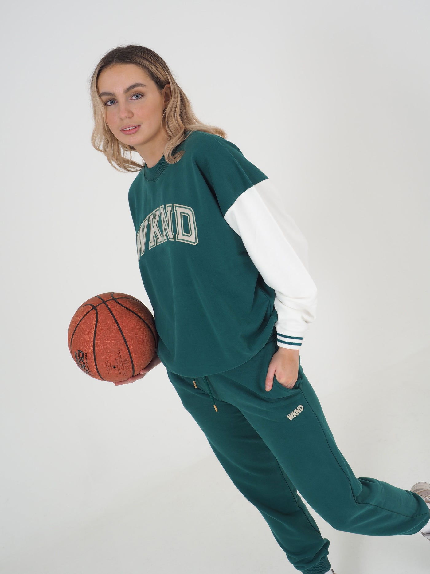 Model with blonde hair wearing a green college varsity style sweatshirt and matching joggers.  Sweatshirt has contrast white sleeves and the text reads WKND. Model is holding a basketball.