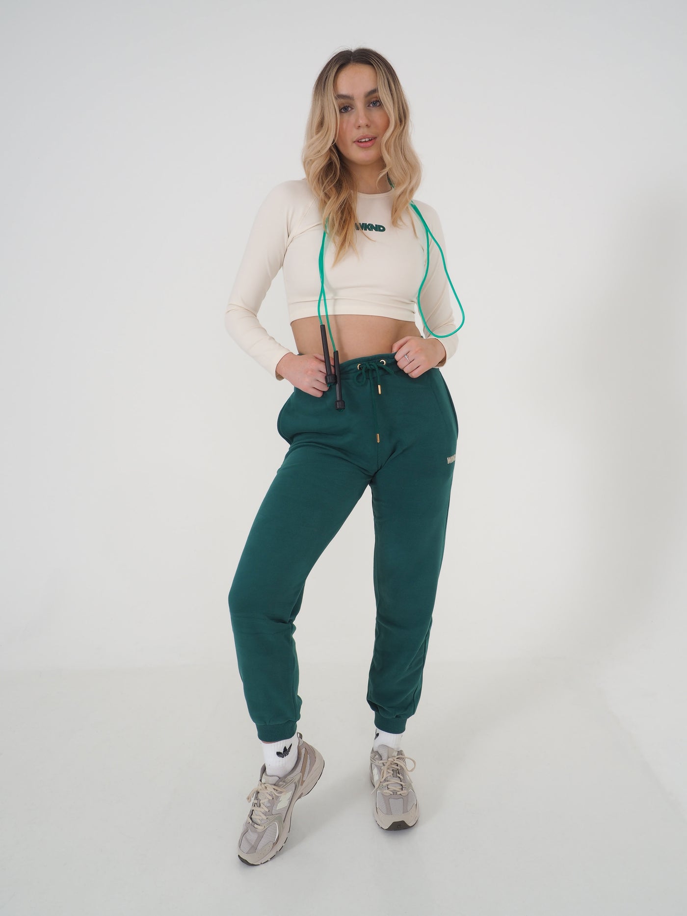 Blonde model is wearing a long sleeve crop top in eggnog cream and matching green joggers.  The WKND logo is green and printed centrally to the chest. Model is also posing with a skipping rope.