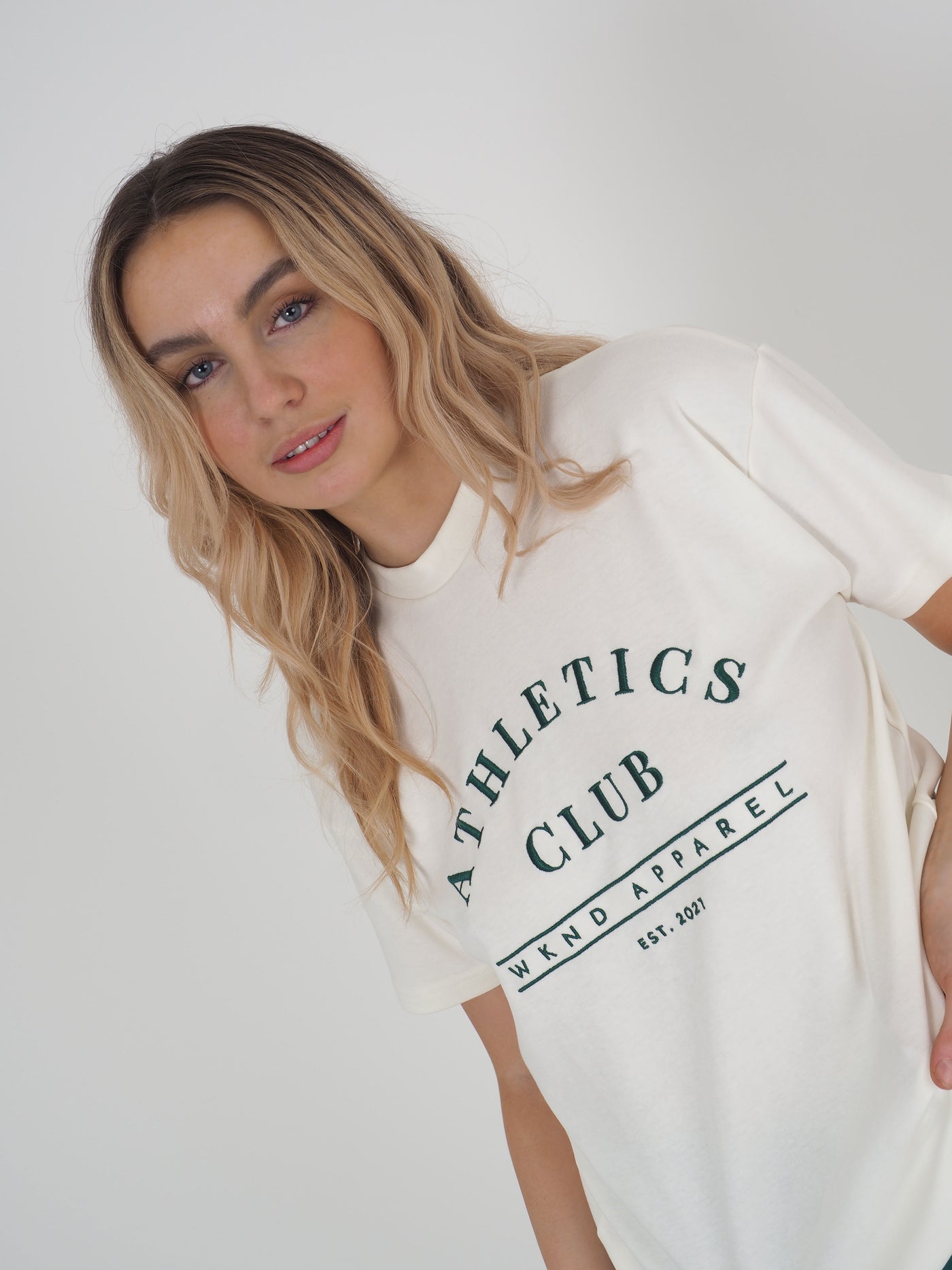 Blonde model wearing a white oversized t-shirt.  T-shirt embroidery spells Athletics Club, WKND Apparel, established 2021.
