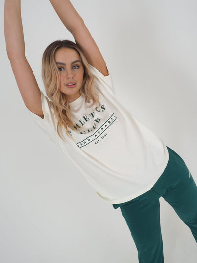 Blonde model has arms in the air and is wearing a white oversized t-shirt and matching green joggers.  T-shirt embroidery spells Athletics Club, WKND Apparel, established 2021.