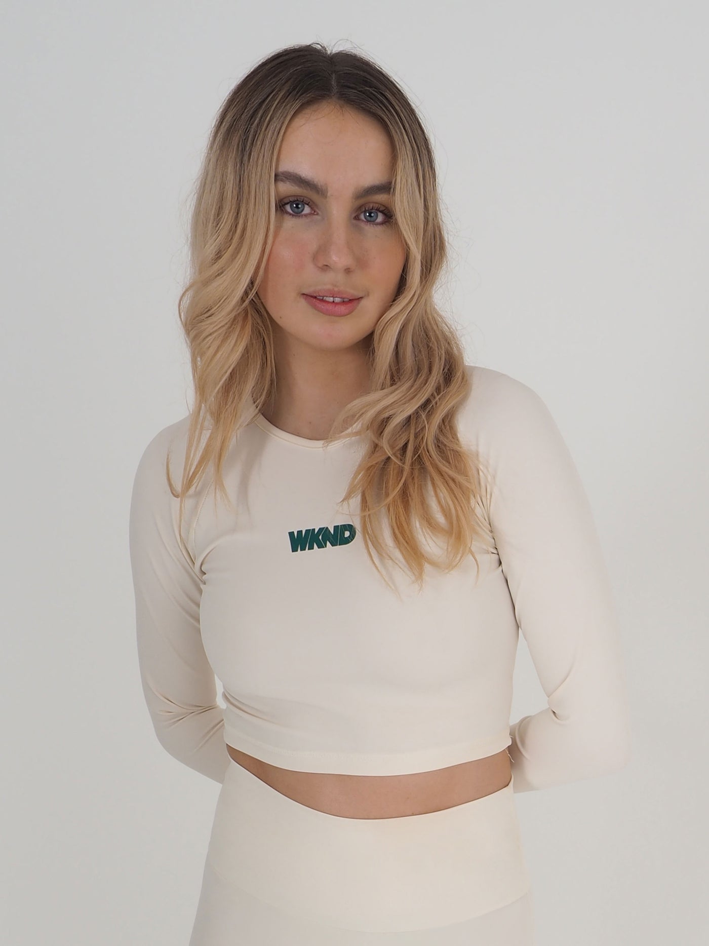 Blonde model is wearing a long sleeve crop top in eggnog cream.  The WKND logo is green and printed centrally to the chest.