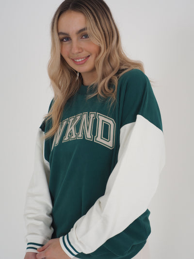 Model with blonde hair wearing a green college varsity style sweatshirt.  Sweatshirt has contrast white sleeves and the text reads WKND.