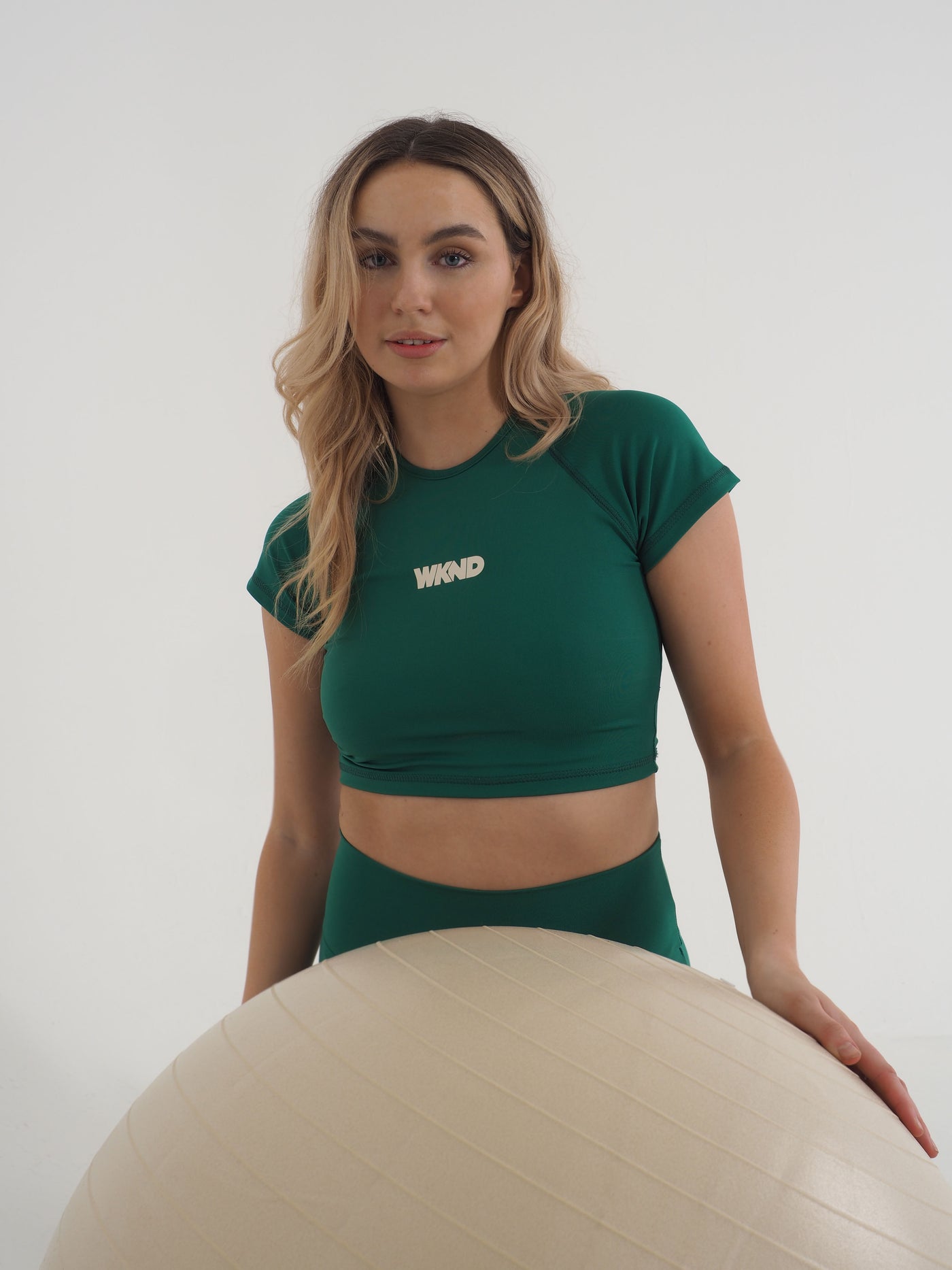 Blonde model wearing a green cropped t-shirt and matching leggings.  The contrast WKND logo is printed centrally on the chest.  Model is posing with a gym yoga ball.