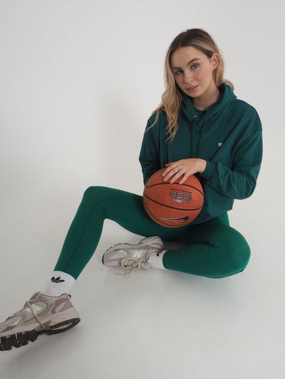 Model with blonde hair is sat on the floor wearing a green hoodie and matching green leggings. WKND Embroidery to the chest. Model is also holding a basketball.