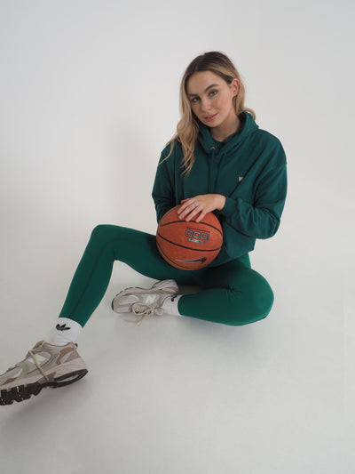 Blonde model is posing on the floor with a basketball.  She is wearing a green hoodie and matching leggings.