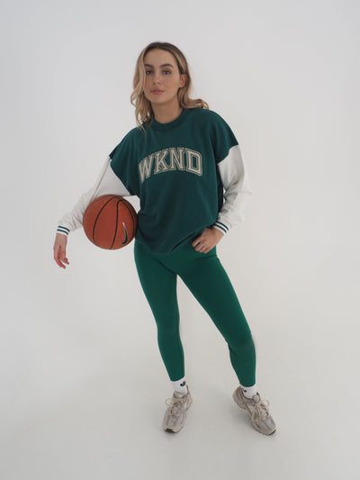 Model with blonde hair wearing a green college varsity style sweatshirt and matching green leggings.  Sweatshirt has contrast white sleeves and the text reads WKND. Model is holding a basketball.