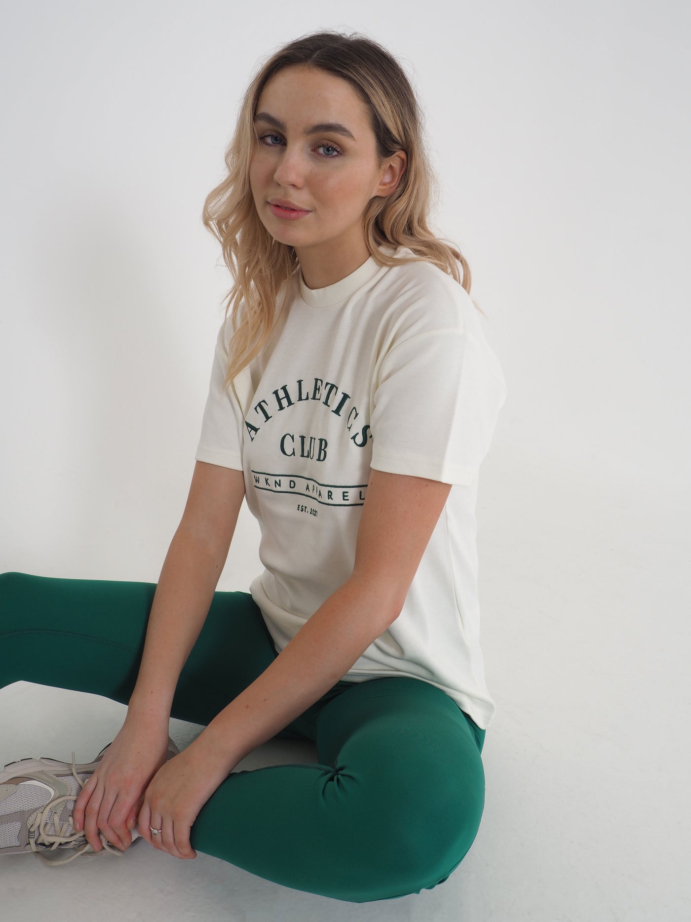 Blonde model is sat on the floor, wearing a white oversized t-shirt and matching green leggings.  T-shirt embroidery spells Athletics Club, WKND Apparel, established 2021.