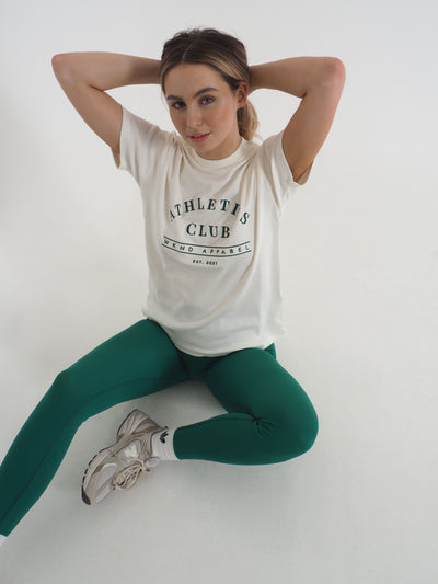 Blonde model is sat on the floor and wearing a white oversized t-shirt and matching green leggings.  T-shirt embroidery spells Athletics Club, WKND Apparel, established 2021.